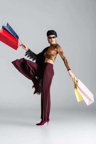 Piena lunghezza della giovane donna alla moda in occhiali da sole e basco che tiene borse della spesa mentre posa sul grigio — Foto stock