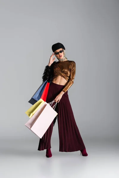 Toute la longueur de la jeune femme à la mode ajuster les lunettes de soleil et tenant des sacs à provisions tout en posant sur le gris — Photo de stock