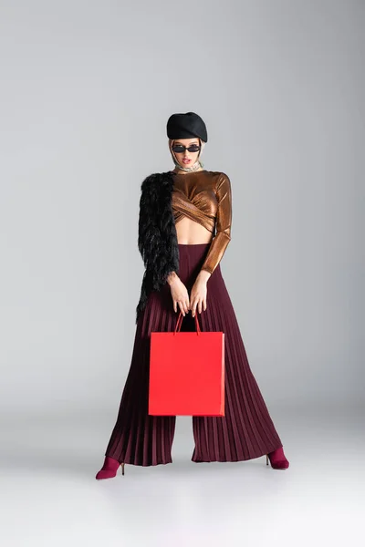 In voller Länge junge Frau im trendigen Outfit mit roter Einkaufstasche, während sie auf grau posiert — Stockfoto
