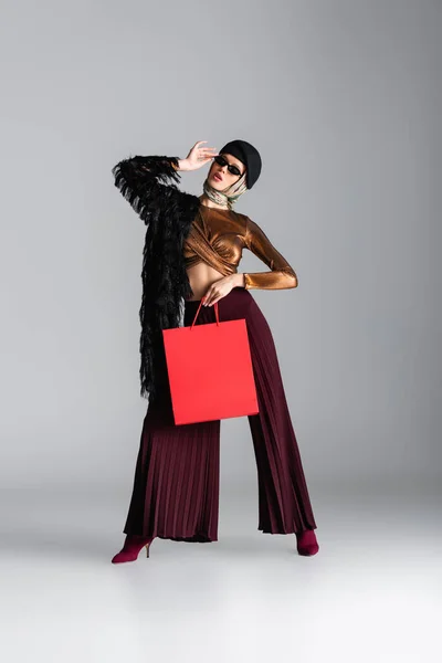 Full length of woman in trendy outfit holding red shopping bag while posing on grey — Stock Photo