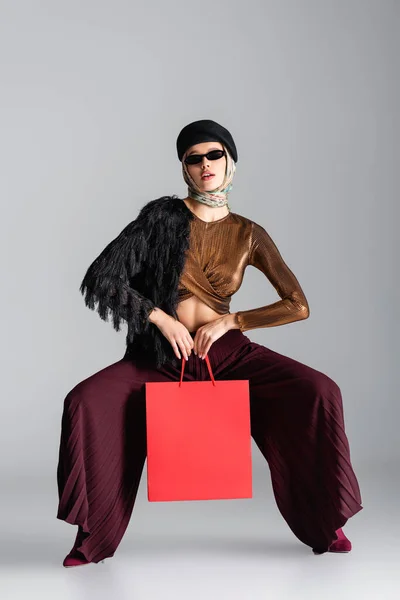 На всю длину молодая женщина в модном наряде держит сумку с покупками, позируя на серой — стоковое фото