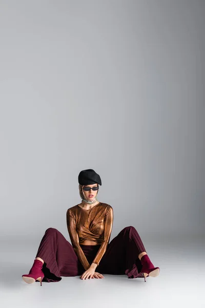 Longitud completa de la mujer con estilo en traje de moda, gafas de sol y boina sentado mientras posa en gris - foto de stock