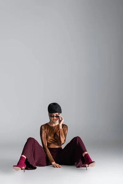 Full length of stylish woman in trendy outfit adjusting sunglasses and sitting while posing on grey — Stock Photo