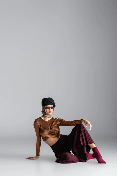 Full length of stylish woman in trendy outfit and sunglasses sitting while posing on grey — Stock Photo