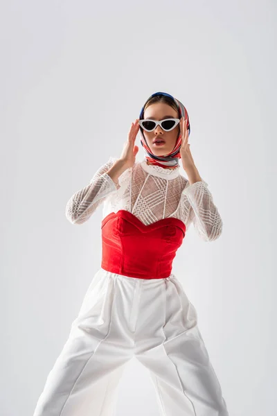Mujer joven y elegante en pañuelo para la cabeza ajustando gafas de sol de moda aisladas en blanco - foto de stock