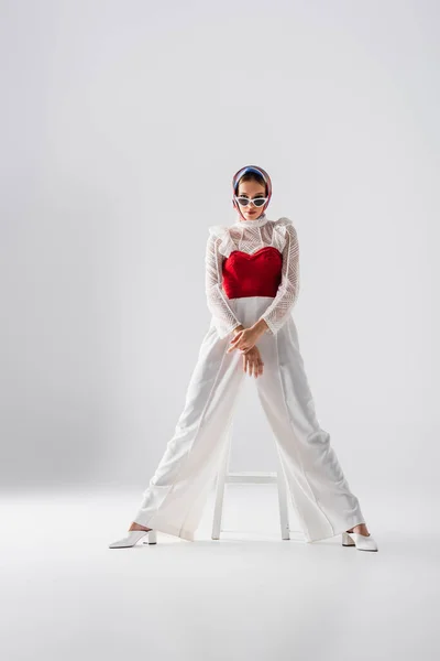 Longitud completa de la mujer joven con estilo en el pañuelo para la cabeza y gafas de sol sentado en el taburete y posando en blanco - foto de stock