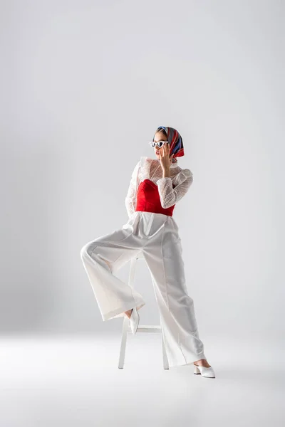 Full length of young woman in headscarf and stylish sunglasses sitting on stool and posing on white — Stock Photo