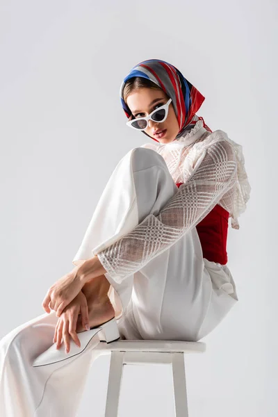 Mujer joven con estilo en pañuelo para la cabeza y gafas de sol sentado en el taburete mientras posa aislado en blanco - foto de stock