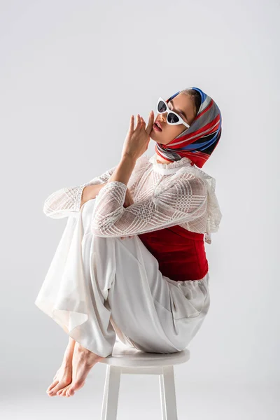 Jeune femme à la mode en foulard et lunettes de soleil assis sur tabouret tout en posant isolé sur blanc — Photo de stock