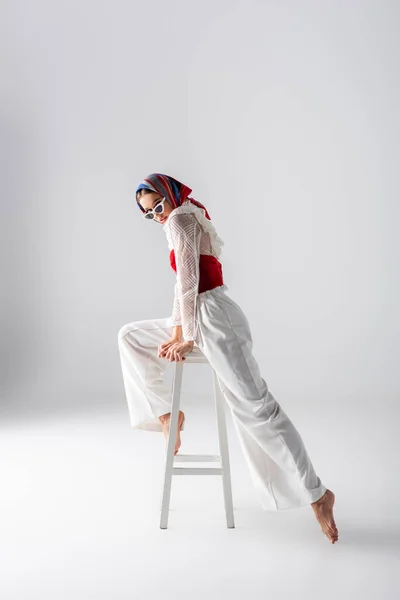 Pleine longueur de femme élégante en foulard et lunettes de soleil posant sur tabouret tout en regardant la caméra sur blanc — Photo de stock