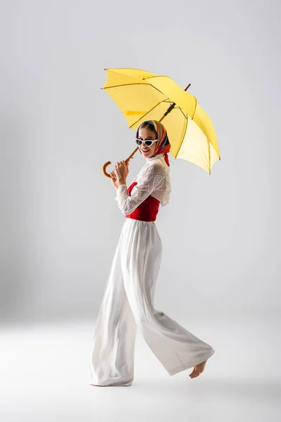 Full length of happy woman in headscarf and sunglasses holding yellow umbrella while posing on white — Stock Photo