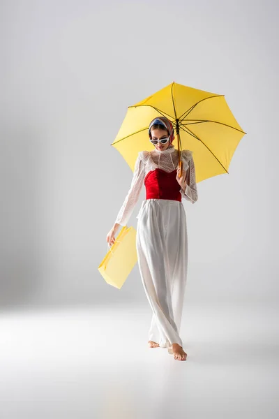 In voller Länge stylische Frau mit Kopftuch und Sonnenbrille, die gelben Regenschirm und Einkaufstasche hält, während sie auf weiß posiert — Stockfoto