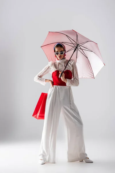 Повна довжина стильної жінки в окулярах і сонцезахисних окулярах тримає червону парасольку і сумку для покупок, позуючи на білому — стокове фото
