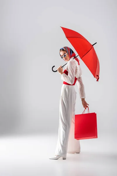 Повна довжина молодої жінки в окулярах і сонцезахисних окулярах тримає червону парасольку і сумку, позуючи на білому — стокове фото