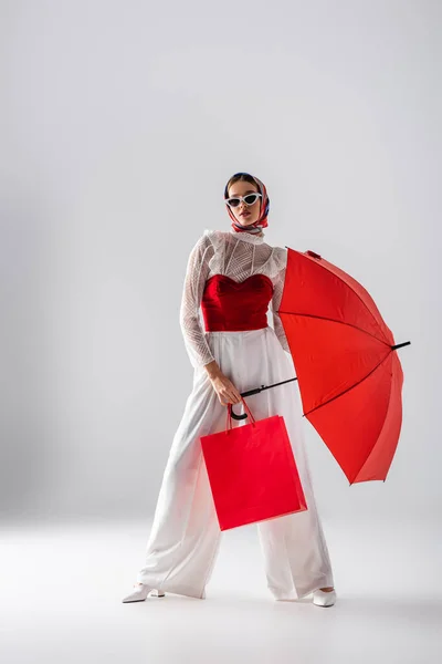 Longitud completa de la mujer de moda en pañuelo para la cabeza y gafas de sol con paraguas rojo y bolsa de compras mientras posa en blanco - foto de stock