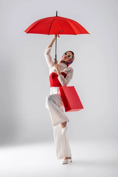 Toute la longueur de la jeune femme en foulard et des lunettes de soleil élégantes tenant parapluie rouge et sac à provisions tout en posant sur blanc — Photo de stock