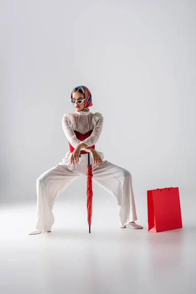 Longitud completa de la mujer con estilo en el pañuelo para la cabeza y gafas de sol sosteniendo paraguas rojo cerca de la bolsa de compras mientras posando en blanco - foto de stock