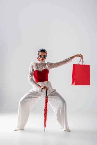 Comprimento total da mulher em lenço de cabeça e óculos de sol na moda segurando guarda-chuva vermelho e saco de compras enquanto posando no branco — Fotografia de Stock
