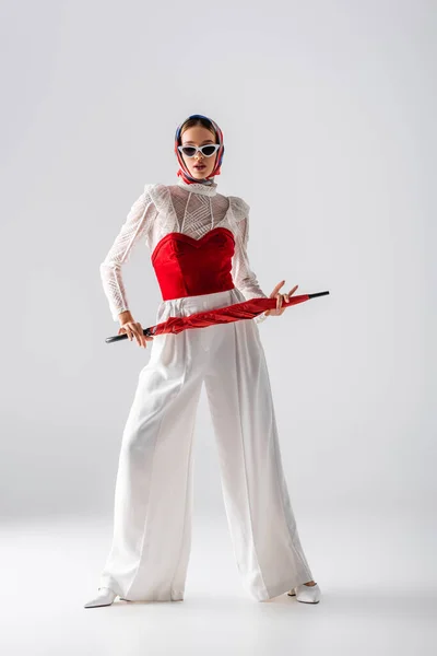 Pleine longueur de femme élégante en foulard et lunettes de soleil tenant parapluie rouge tout en posant sur blanc — Photo de stock