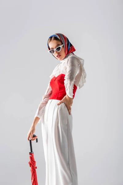 Elegante donna in velo e occhiali da sole con in mano ombrello rosso mentre posa su bianco — Foto stock