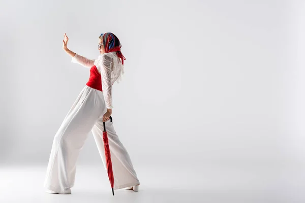 Comprimento total da mulher elegante em lenço de cabeça e óculos de sol segurando guarda-chuva vermelho enquanto gesticulando em branco — Fotografia de Stock