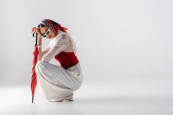 Full length of stylish woman in headscarf and sunglasses holding red umbrella while sitting on white — Stock Photo