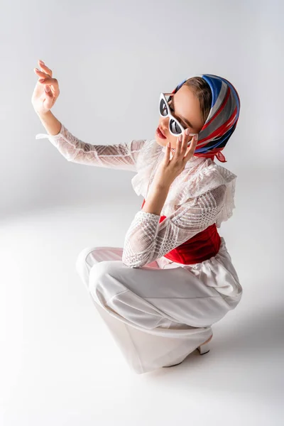 Stylish woman in headscarf adjusting sunglasses, gesturing and sitting on white — Stock Photo