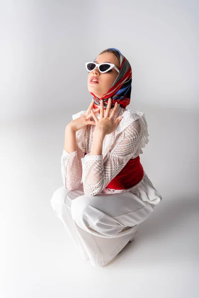 Femme élégante en foulard et lunettes de soleil assis sur blanc — Photo de stock