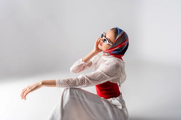 Mujer de moda en pañuelo para la cabeza ajustando gafas de sol y sentado en blanco - foto de stock