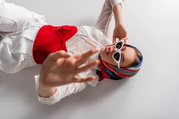 Draufsicht der trendigen Frau mit Kopftuch und Sonnenbrille, die mit ausgestreckter Hand auf Weiß liegt — Stockfoto