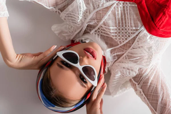 Vue de dessus de la femme à la mode dans les lunettes de soleil et le foulard couché tout en regardant la caméra sur blanc — Photo de stock