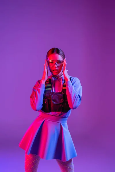 Young woman adjusting sunglasses and looking at camera on purple — Stock Photo