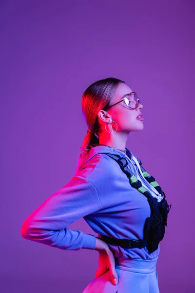 Вид сбоку стильной женщины в солнцезащитных очках, стоящей с рукой на бедре, изолированной на фиолетовом — стоковое фото