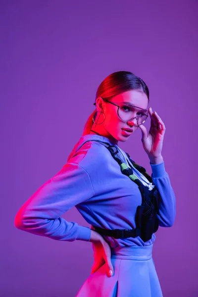 Stylish woman adjusting sunglasses and standing with hand on hip isolated on purple — Stock Photo