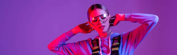 Mujer con estilo en gafas de sol posando aislado en púrpura con iluminación rosa, pancarta — Stock Photo