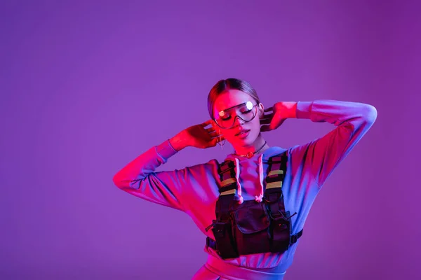 Стильна жінка в сонцезахисних окулярах позує ізольовано на фіолетовому з рожевим освітленням і копіюванням простору — стокове фото