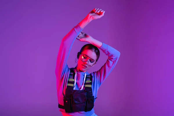 Stylish woman in sunglasses with hands above head posing on purple — Stock Photo