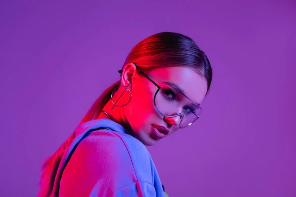 Молодая стильная женщина в солнечных очках, смотрящая на камеру, изолированную на фиолетовый — стоковое фото