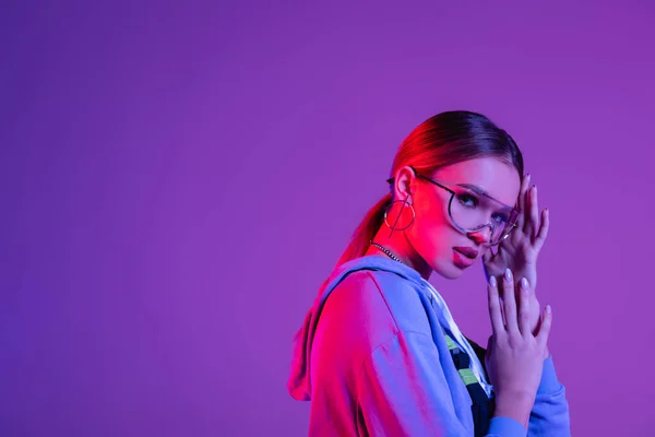 Jeune femme à la mode dans les lunettes de soleil en regardant la caméra isolée sur violet — Photo de stock