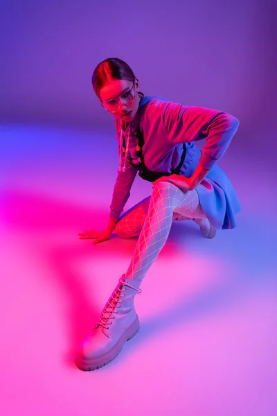 Jeune femme élégante dans des lunettes de soleil et tenue à la mode assis tout en regardant loin sur violet et rose — Photo de stock