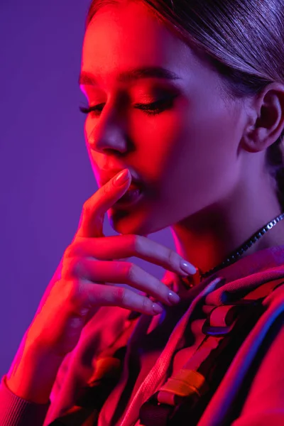 Iluminación rosa en la cara de la joven mujer pensativa tocando los labios aislados en púrpura - foto de stock