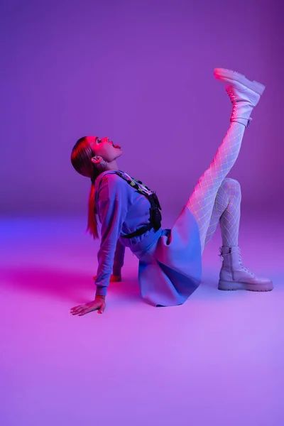 Full length of stylish woman in trendy outfit sitting and sticking out tongue on purple — Stock Photo