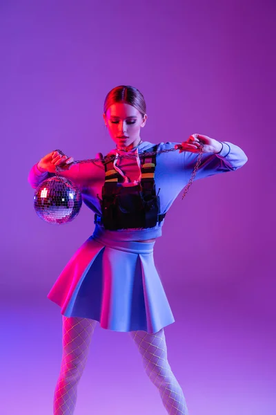 Jovem e elegante mulher na saia posando com bola de discoteca brilhante em roxo — Fotografia de Stock