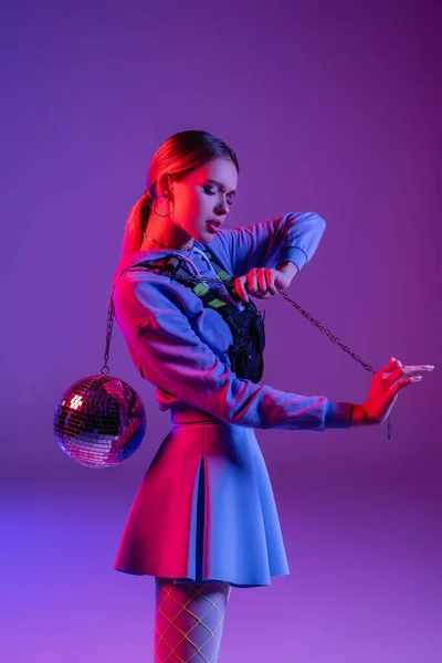 Jovem e na moda mulher posando com bola de discoteca brilhante em roxo — Fotografia de Stock