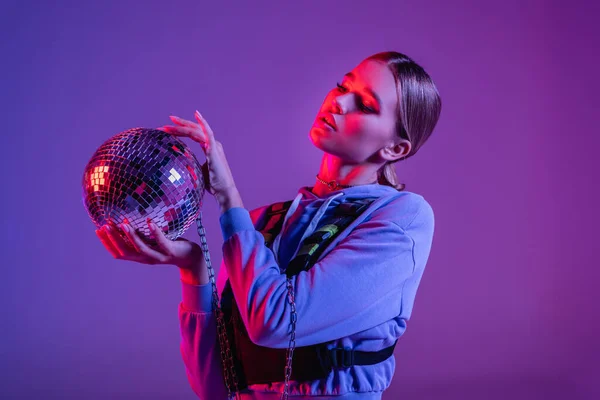 Jovem e elegante mulher olhando para bola de discoteca brilhante em roxo — Fotografia de Stock