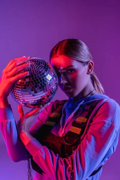 Молода і стильна жінка тримає блискучий диско м'яч і дивиться на камеру ізольовано на фіолетовому — стокове фото