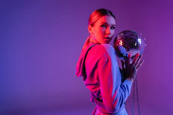 Jovem e elegante mulher segurando bola de discoteca brilhante e olhando para longe em roxo — Fotografia de Stock
