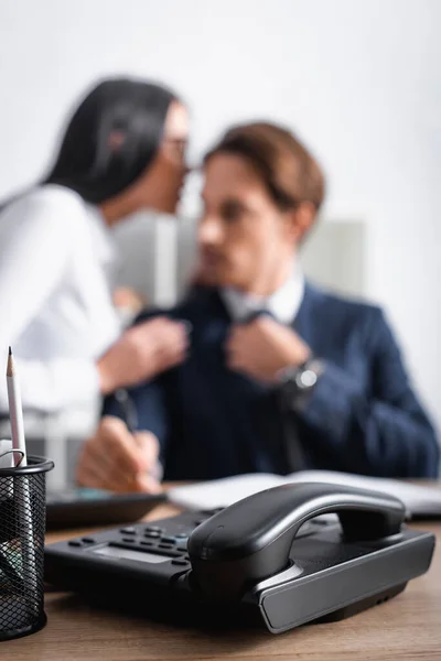 Selective focus of landline phone near businesswoman flirting with colleague in office on blurred background — Stock Photo