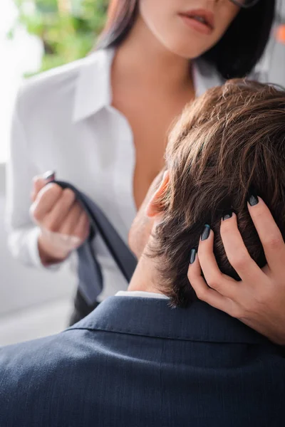 Vista ritagliata della segretaria seducente tirando cravatta e toccando la testa dell'uomo d'affari in ufficio, sfondo sfocato — Foto stock