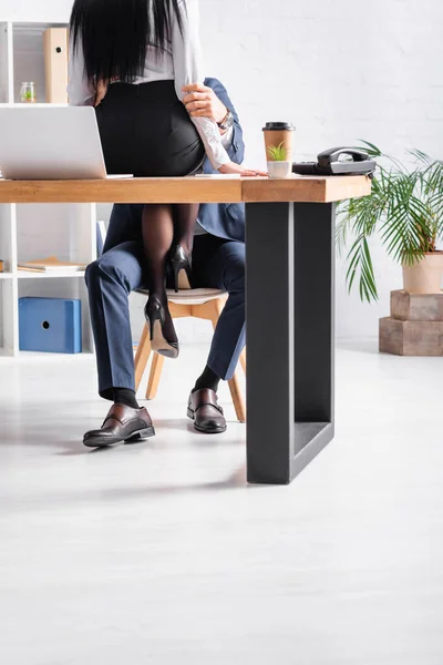 Cropped view of businesswoman sitting on desk while seducing colleague in office — Stock Photo